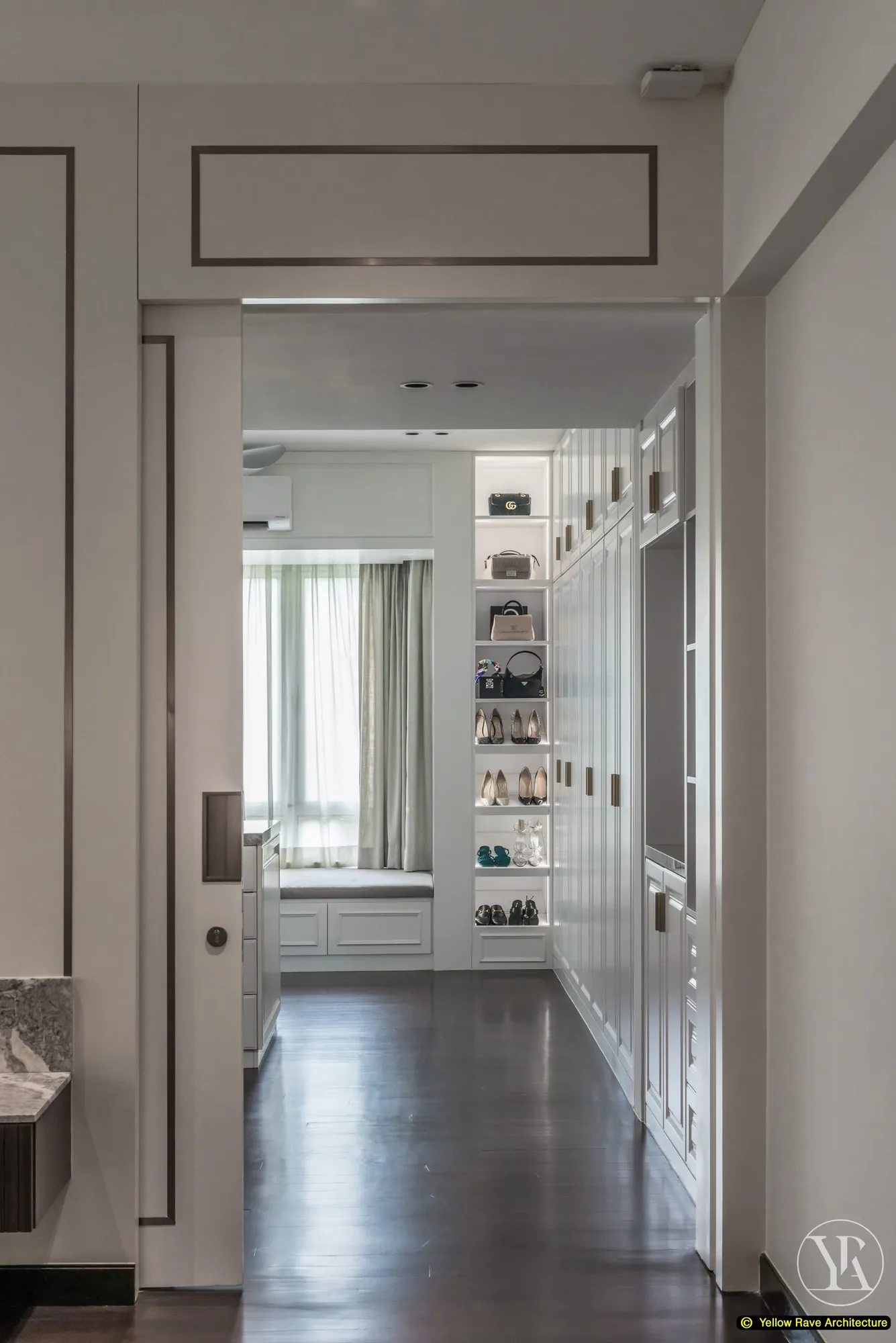 Pristine Walk-in Closet — view from entry showing white panel-moulded cabinets lining both walls, central island and window seat with open shelving visible at the far end