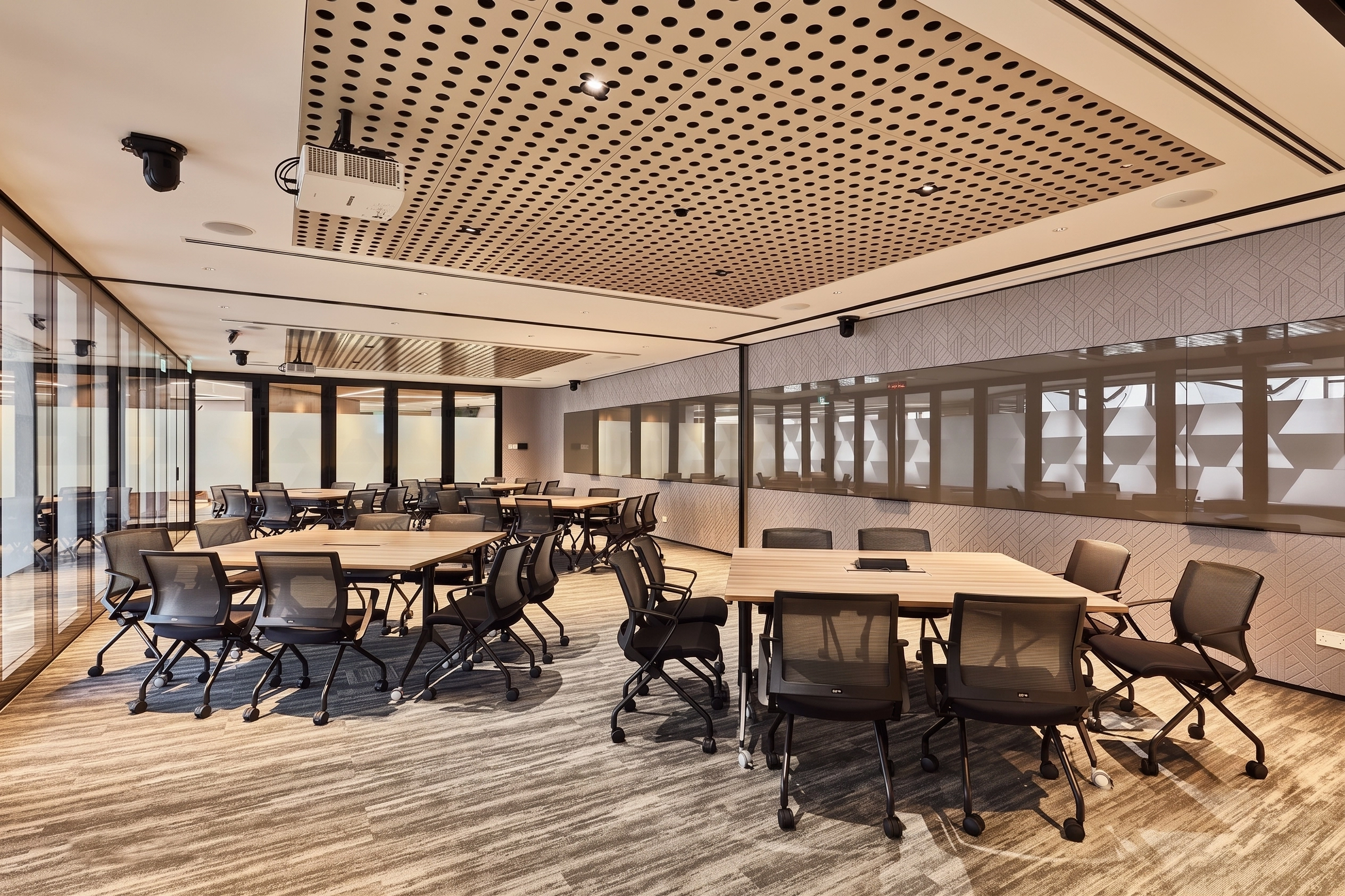 Meeting room with two timber tables, black mesh chairs, a perforated ceiling, and patterned glass partitions